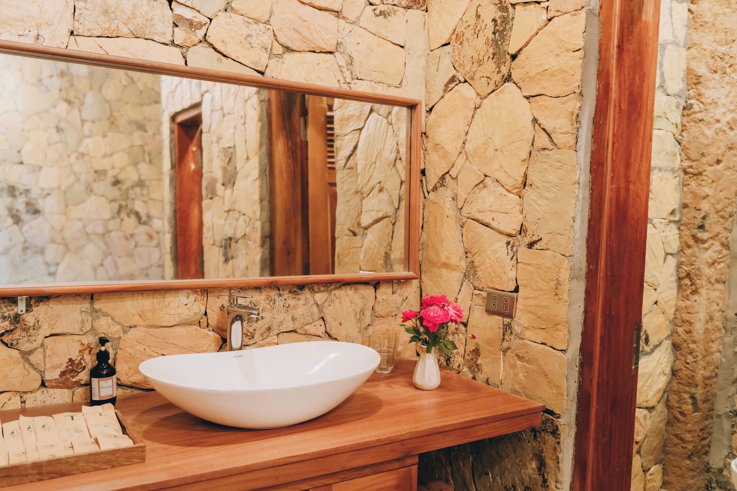 Une salle de bain avec un mur en pierre, un meuble-lavabo en bois, un lavabo blanc, un miroir, une porte en bois et un vase rempli de fleurs.
