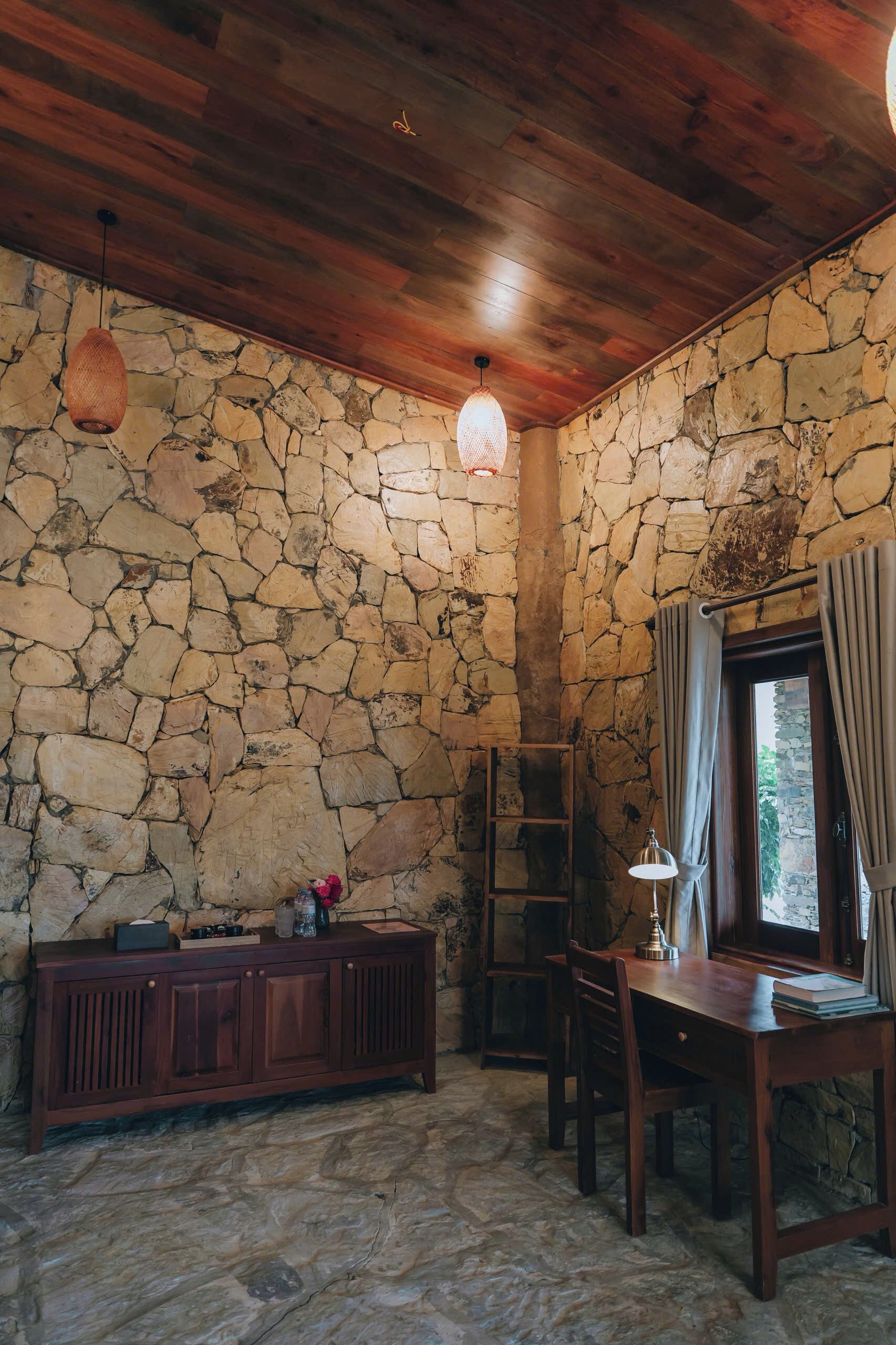 A rustic room with stone walls, wooden ceiling, a wooden table with chairs, a cabinet, and a hanging light fixture.