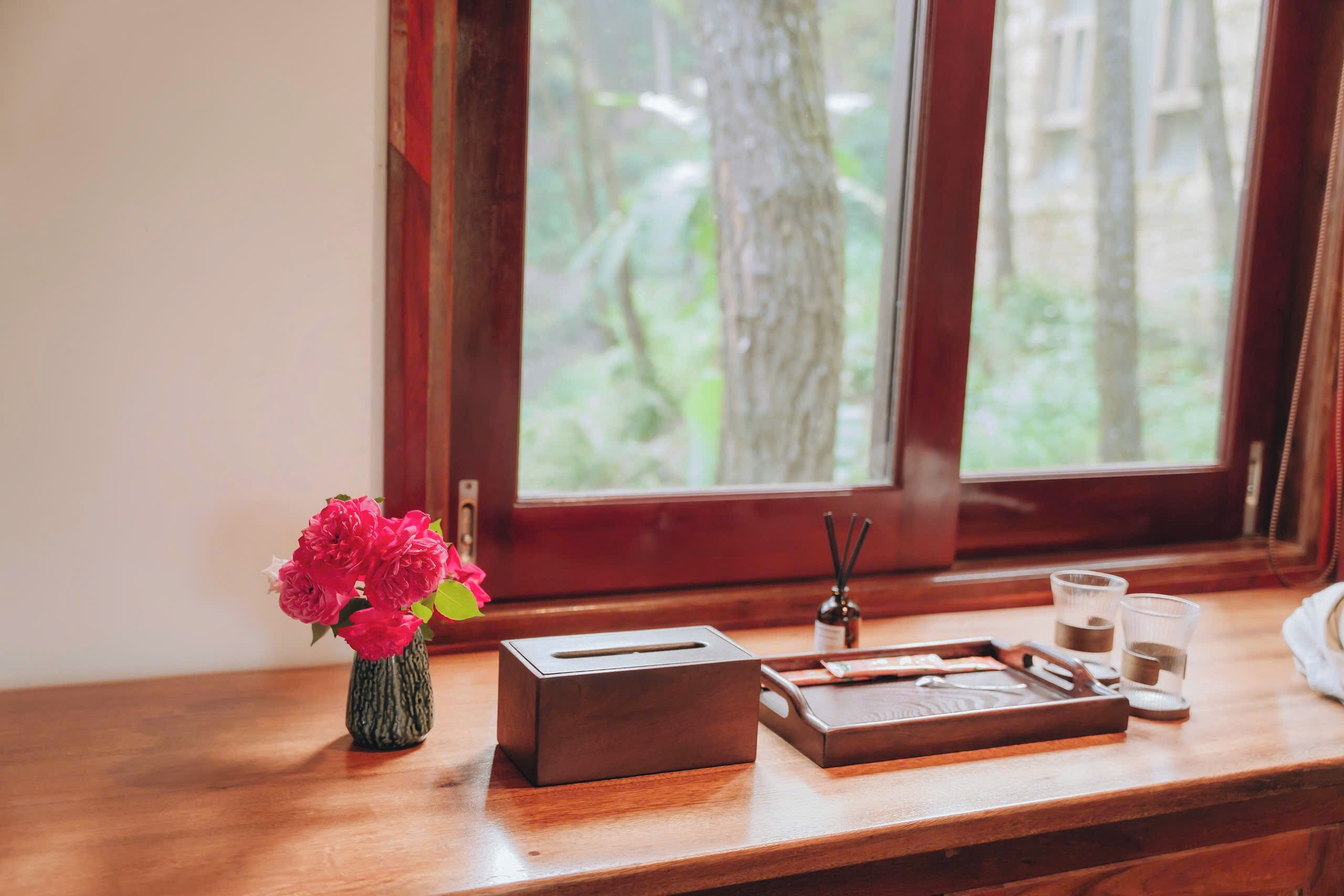 A wooden table with a tissue box, a tray, a vase with flowers, and a window with a view of trees.