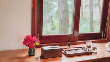 A wooden table with a tissue box, a tray, a vase with flowers, and a window with a view of trees.