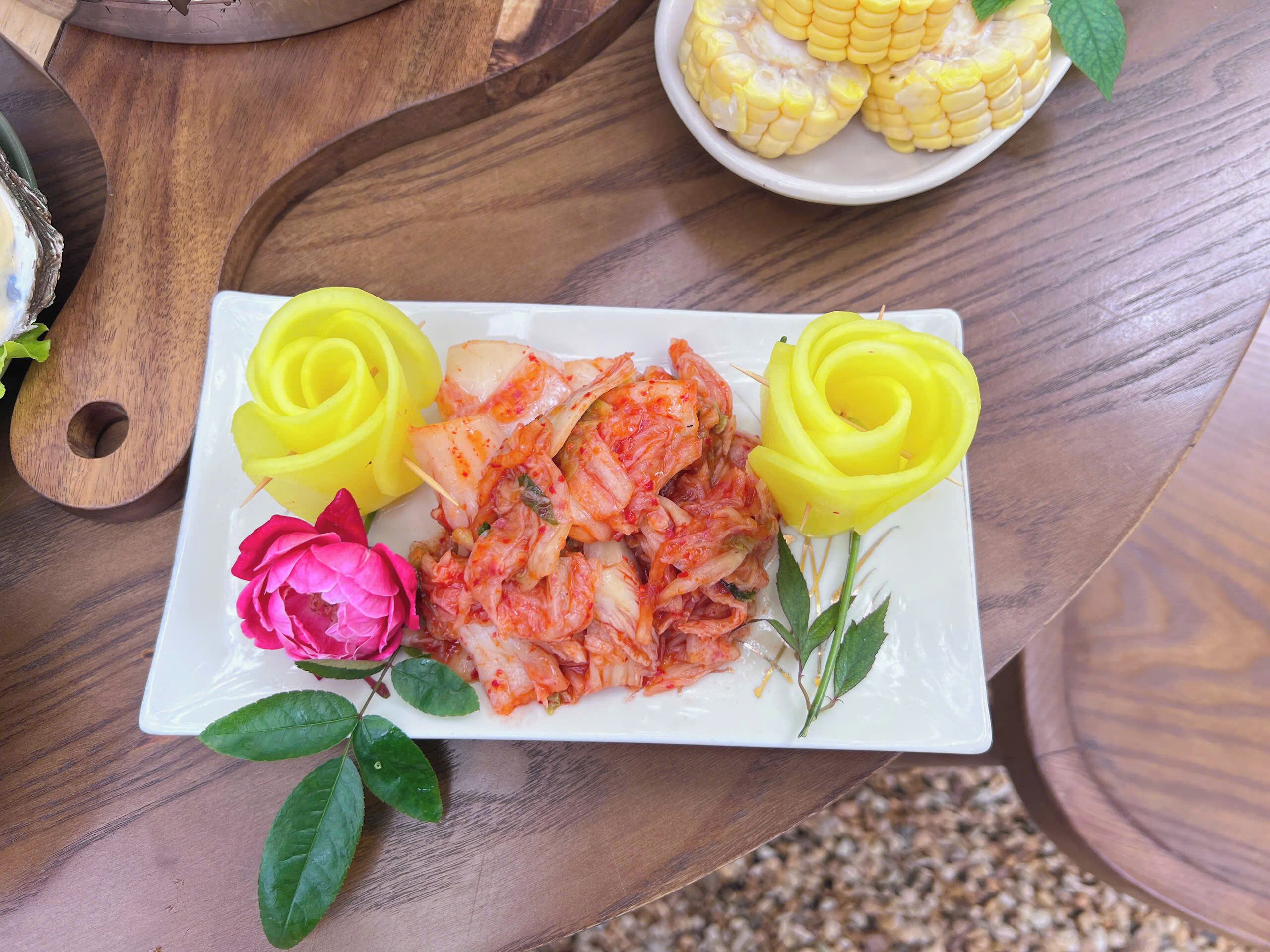 A plate of kimchi with two yellow rose-shaped decorations and a pink flower on a wooden table.