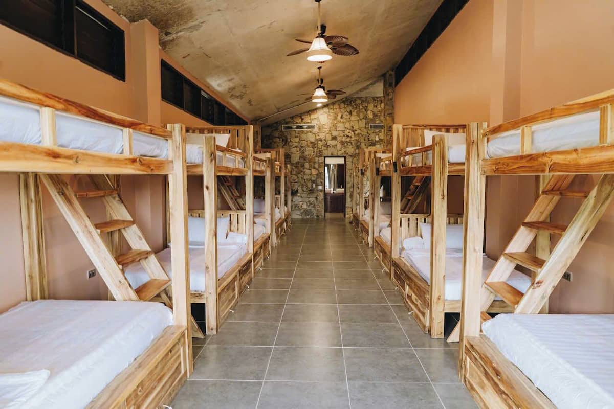 A row of bunk beds with wooden frames and white bedding, arranged in a narrow corridor with a stone wall and a ceiling fan.