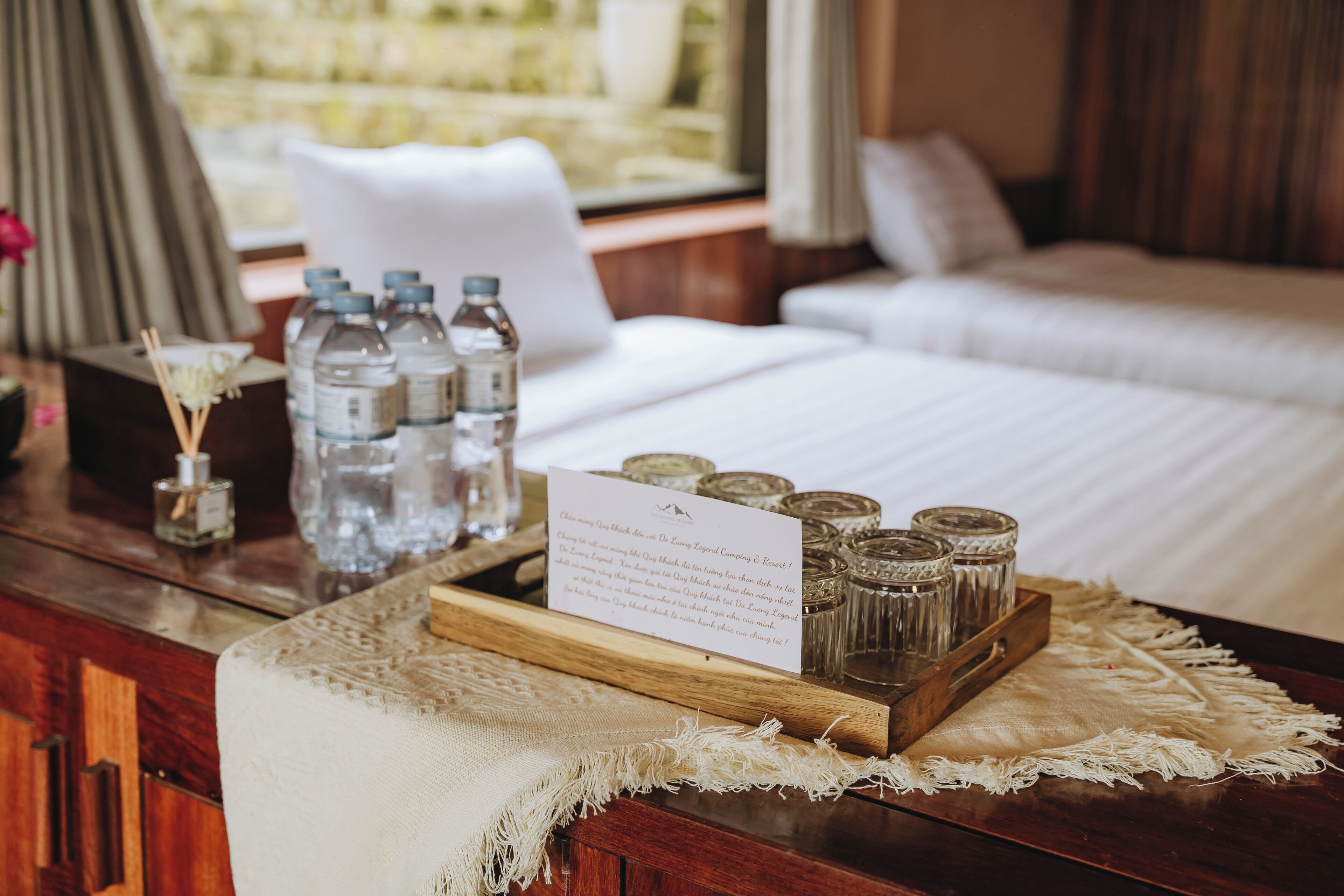A neatly arranged hotel room with a bed, a wooden side table with water bottles, a tray of glass jars, and a note on the table.