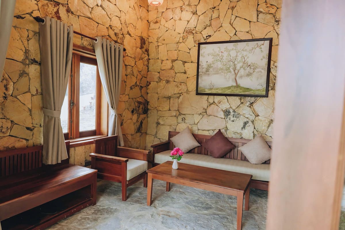 A room with a stone wall, a wooden bench, a sofa with cushions, a wooden coffee table, and a window with curtains.
