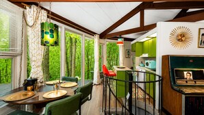 A dining area with a table set for four, a kitchen with green cabinets, and a view of trees through large windows.