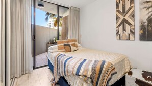 A bedroom with a bed, curtains, a view through a sliding door, and artwork on the wall. - Beachside Studio With Pool and Gym Access (Sydney)