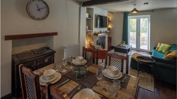 A dining area with a wooden table set for four, a clock on the wall, and a cozy living room with a green sofa and a television. - Stylish & Spacious Retreat in Troedyrhiw | Sleeps 6 | Garden Decking (Troedyrhiw)