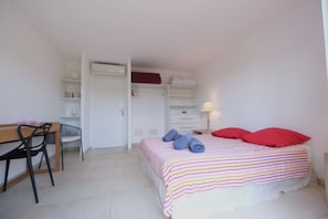 Un dormitorio con cama, escritorio, sillas, armario y mesita de noche con lámpara.
