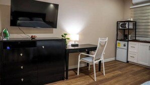 Un espace de vie compact comprenant un téléviseur à écran plat fixé au mur, un bureau avec une chaise, une petite kitchenette équipée d’un four à micro-ondes et un meuble avec des tiroirs.