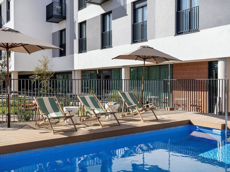 Zona junto a la piscina con sillones de descanso y sombrillas, piscina con azulejos y un edificio con balcones.