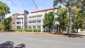 A multi-story building with balconies, a clear sky, and trees along the street. - Relaxed 2br, 2bth Apartment With Balcony & Parking (Canberra)