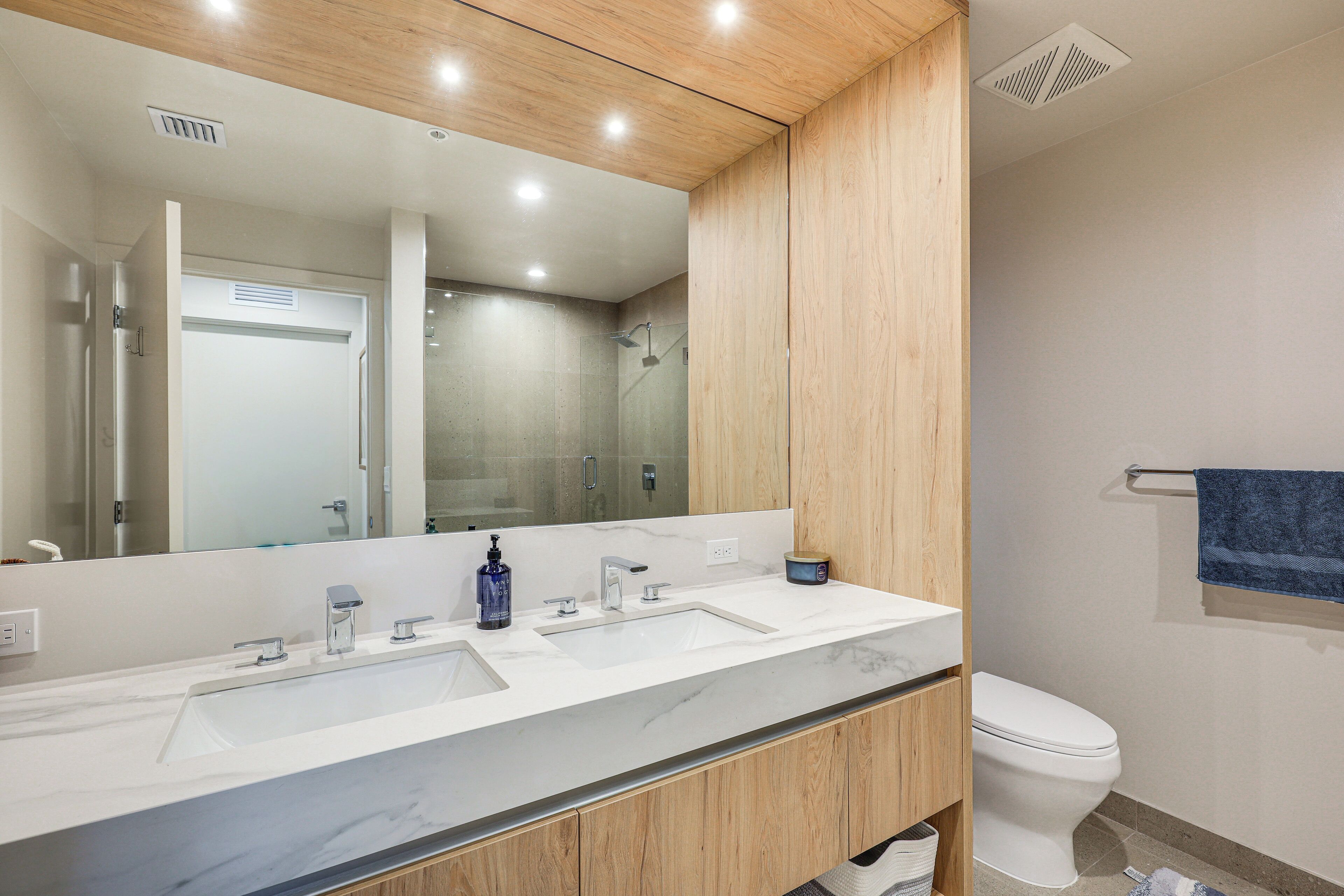 Un baño moderno con un espejo grande, dos lavamanos, un inodoro y armarios de madera.