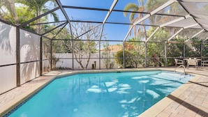 Une piscine intérieure à l’eau d’un bleu limpide, entourée d’une structure en métal et de panneaux de verre, avec des palmiers et un espace patio visibles au fond.