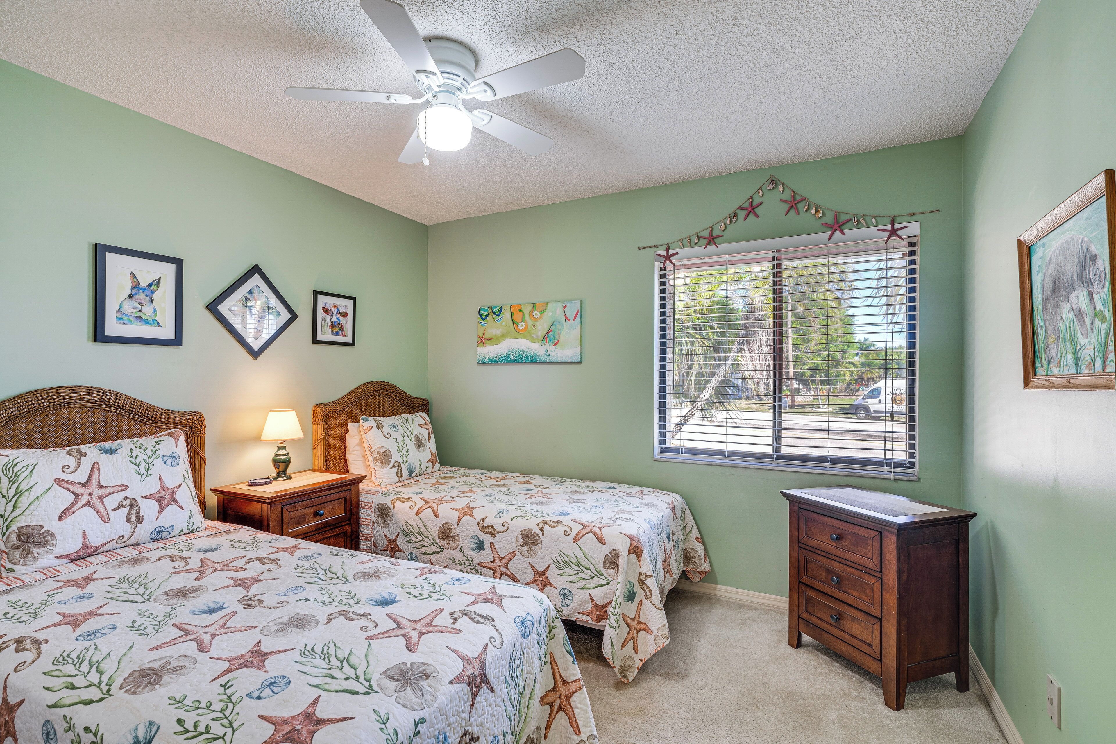A bedroom with two beds, a ceiling fan, a window with blinds, and artwork on the walls.