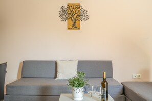 Een woonkamer met een grijze bank, een witte tafel met een plant en wijnkannen, en een wanddecoratie van een boom.