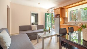 A modern living room with a sofa, a dining table, and a kitchen area with wooden cabinets and a window. - Anna's Two-Bedroom Apartment with Mountain View, Wi-Fi, and Air Conditioning (Νικιάνα)