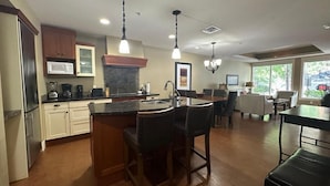 A spacious kitchen with a central island, bar stools, a dining area with a table and chairs, and a view of the outdoors through large windows. - Solara 2BR Retreat Pool- Hot Tub -113 (Canmore)
