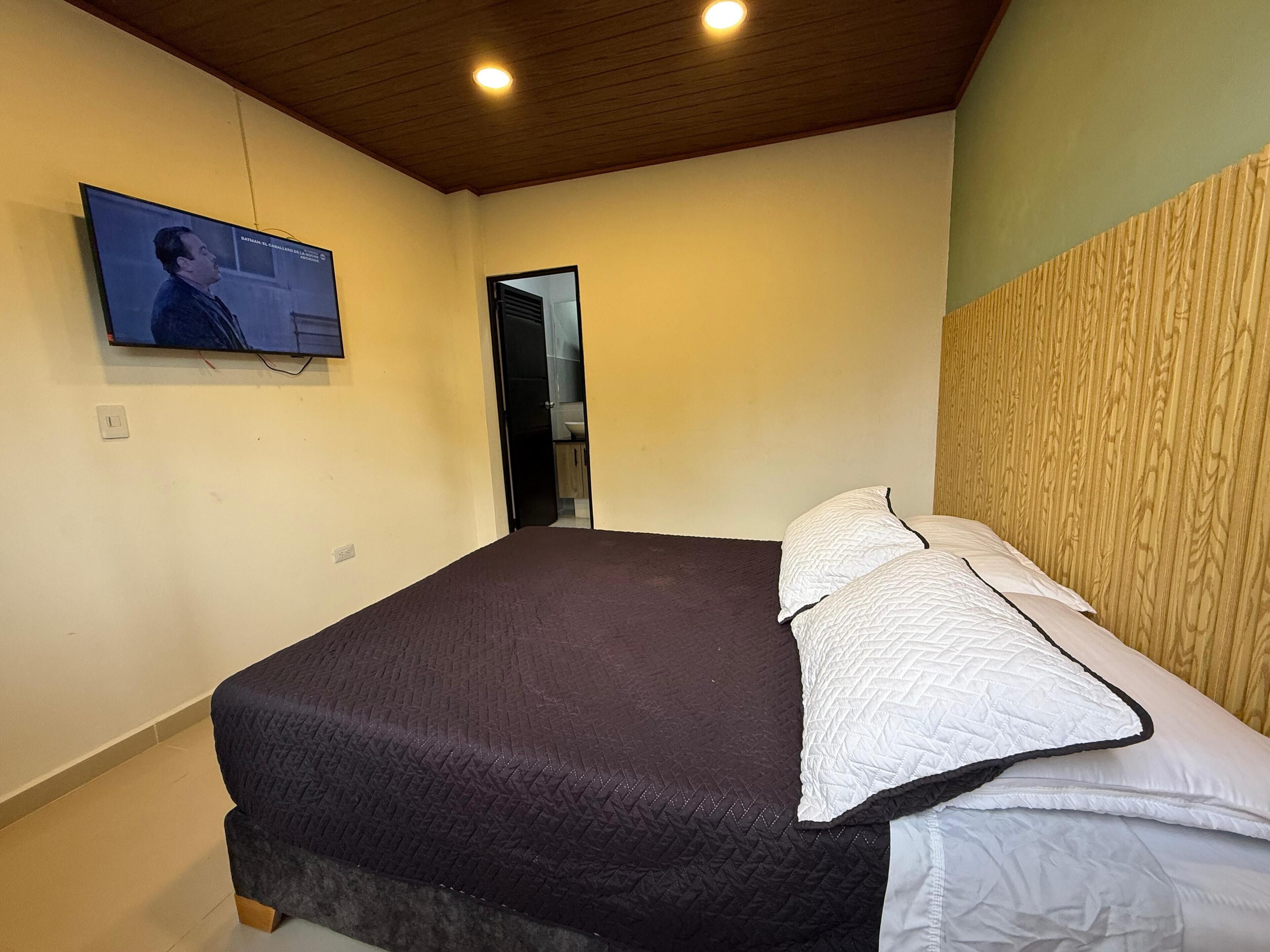 Un dormitorio con una cama grande, cabecera de madera, televisor montado en la pared y una puerta que da al baño.