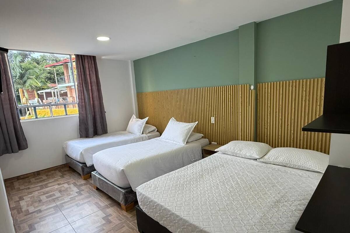 Een hotelkamer met drie bedden, een groot raam met gordijnen en uitzicht op buiten.
