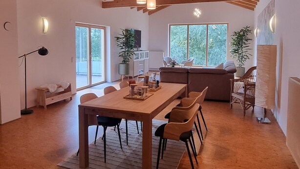 Een ruime eethoek met een houten tafel en stoelen, een hoog plafond met zichtbare balken en grote ramen die voor daglicht zorgen.