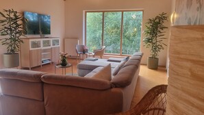 Een moderne woonkamer met een groot raam, een zithoek, een televisie en potplanten.