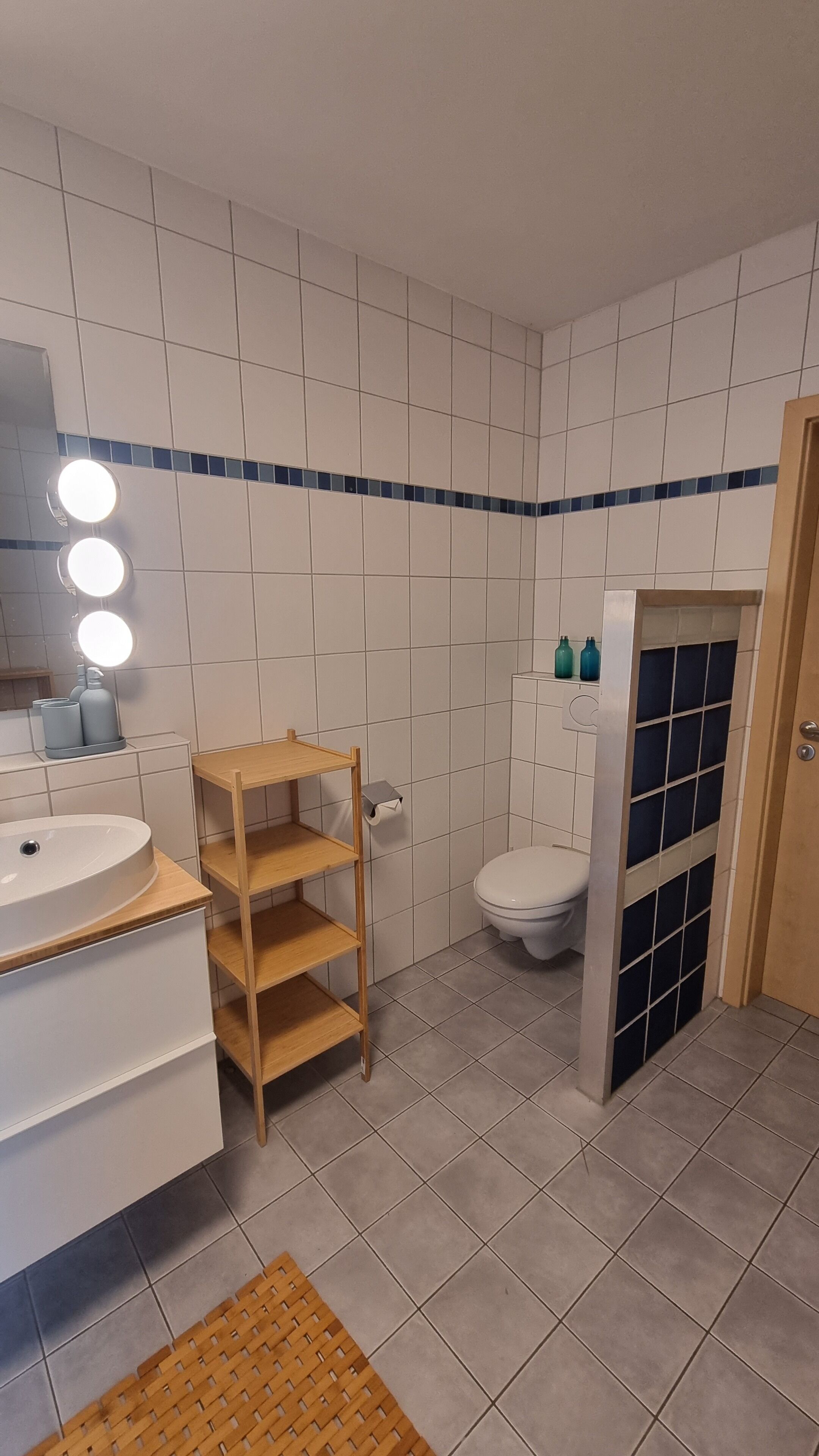 A bathroom with white tiled walls, a wooden vanity with a sink, a toilet, and a shower area with a glass door.