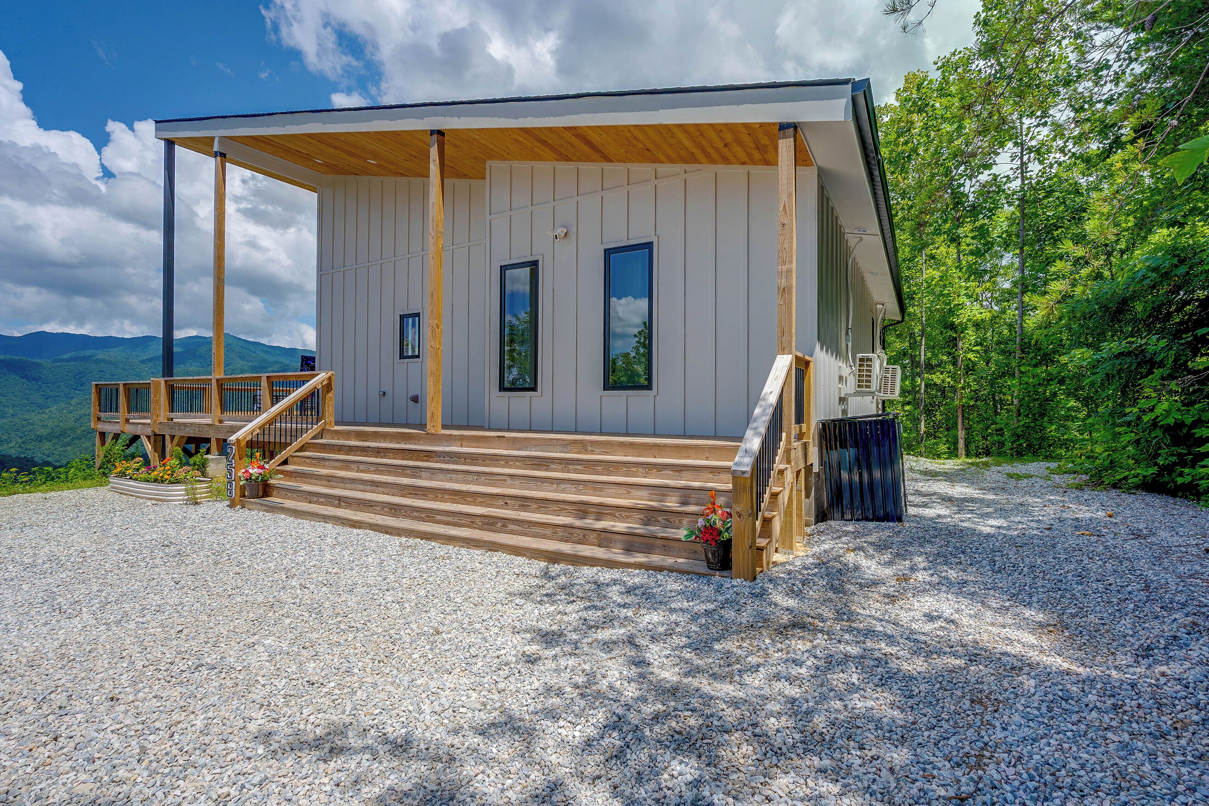 A two-story wooden cabin with a covered porch, a gravel driveway, and a mountain backdrop.