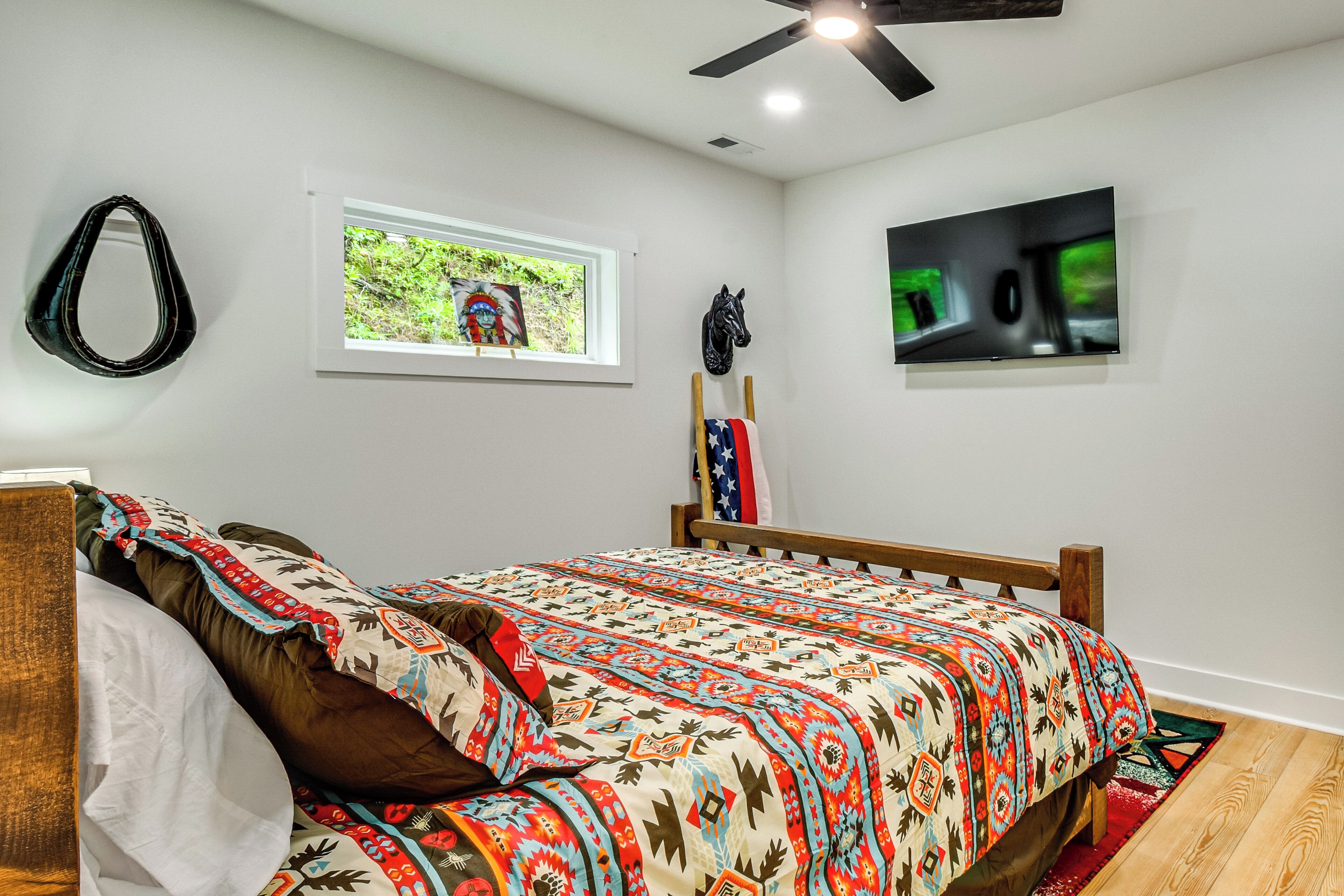 A bedroom with a bed, a ceiling fan, a window with a view of greenery, and a mounted television.
