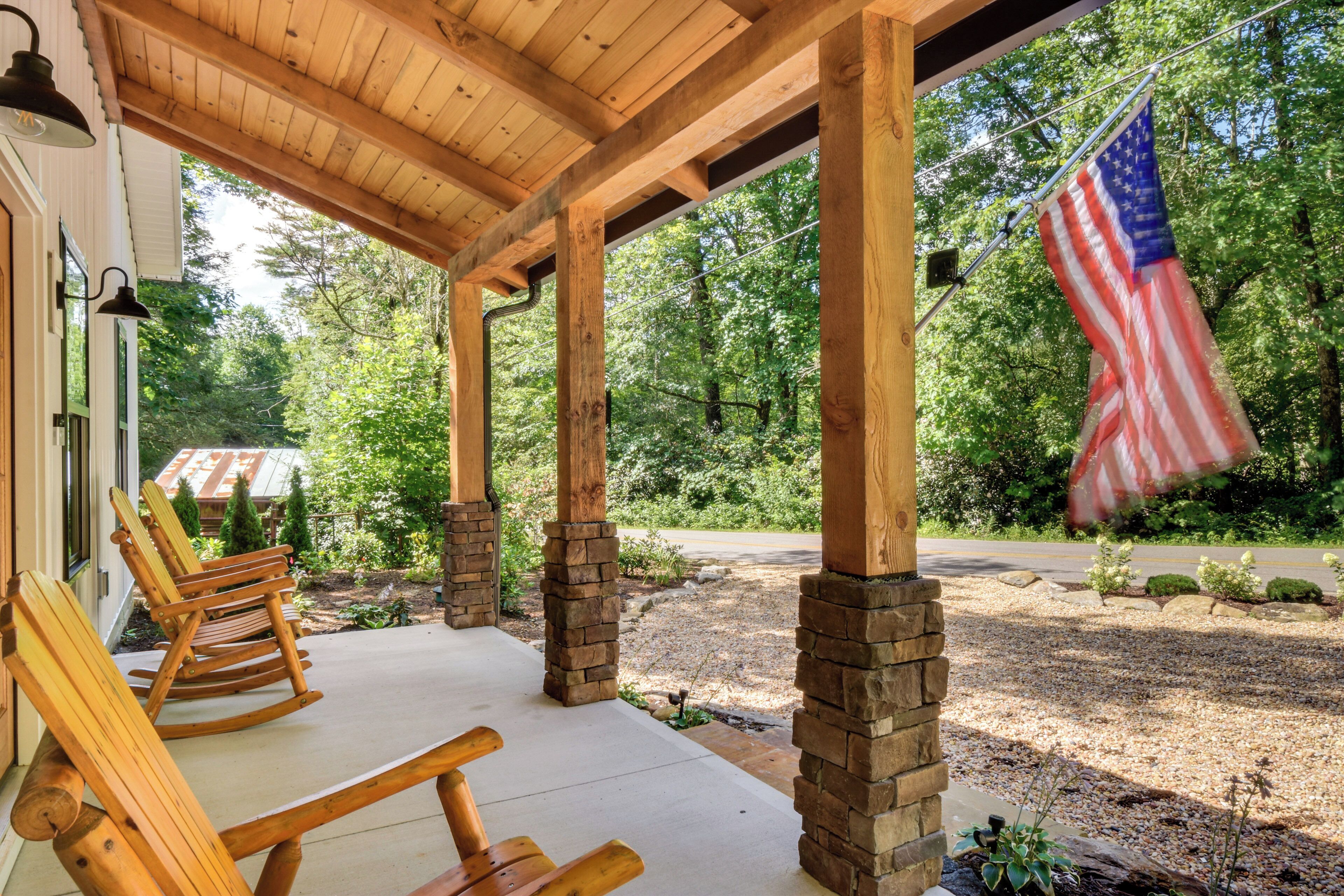 A covered porch with wooden rocking chairs, a flag, and a view of a forested area.