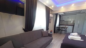 Quarto de hotel moderno com sofá, cama com toalhas, área de refeições e cozinha compacta.