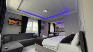 Quarto de hotel moderno com cama, sofá, televisão e cozinha compacta.