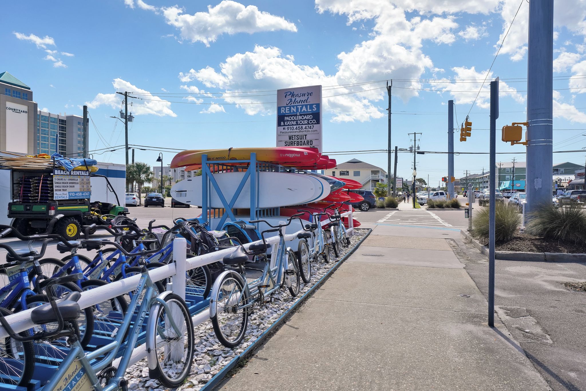 A beach rental facility with kayaks and paddleboards, a sign for 'Pleasure Island Rentals & Paddleboard & Kayak Tour Co.', and a row of bicycles.