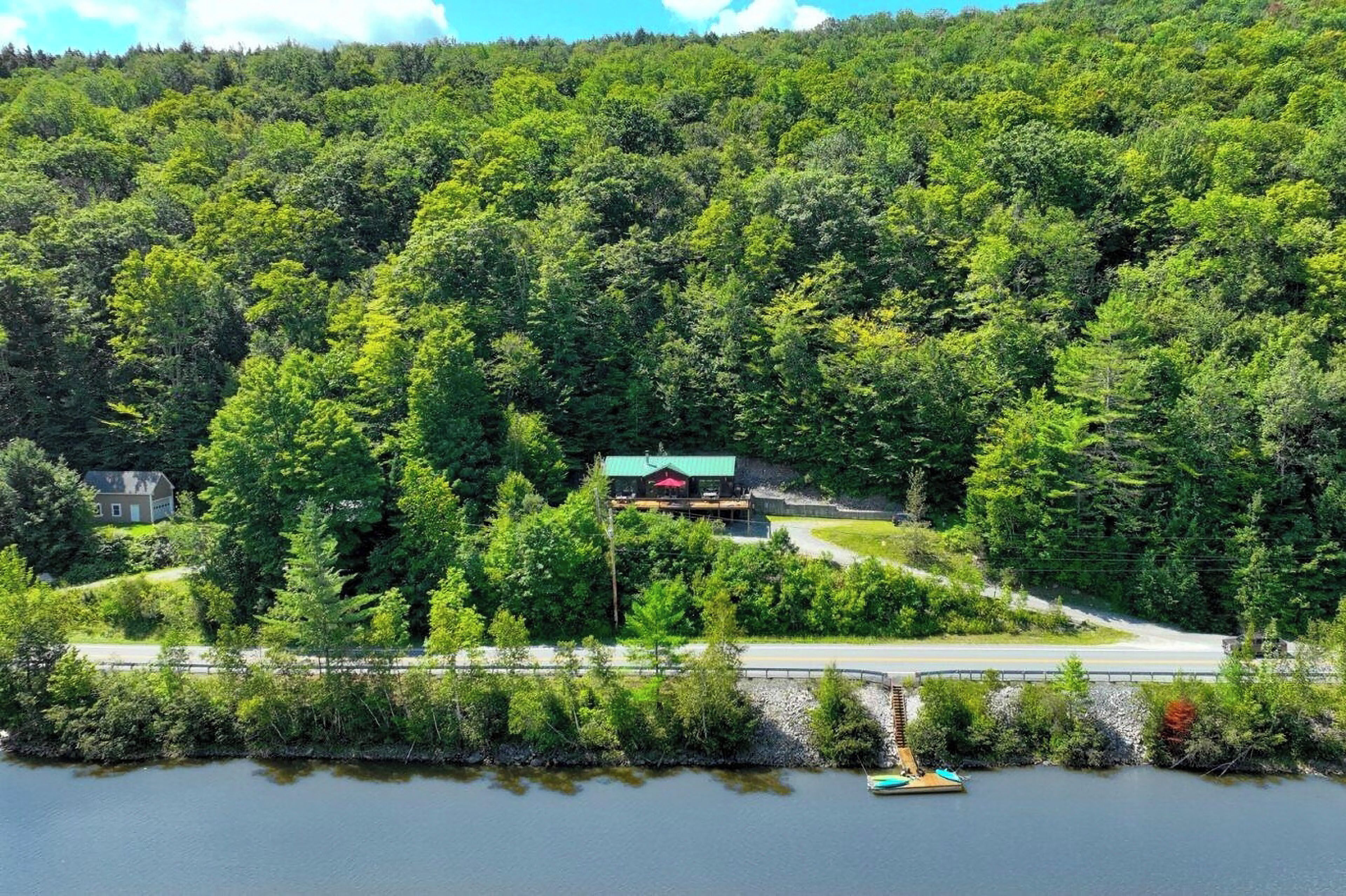 Eine am See gelegene Holzhütte mit rotem Dach, umgeben von dichtem Wald, erreichbar über eine asphaltierte Straße.