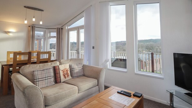 A living room with a sofa, a coffee table, and a TV. There are large windows and a balcony with a view.