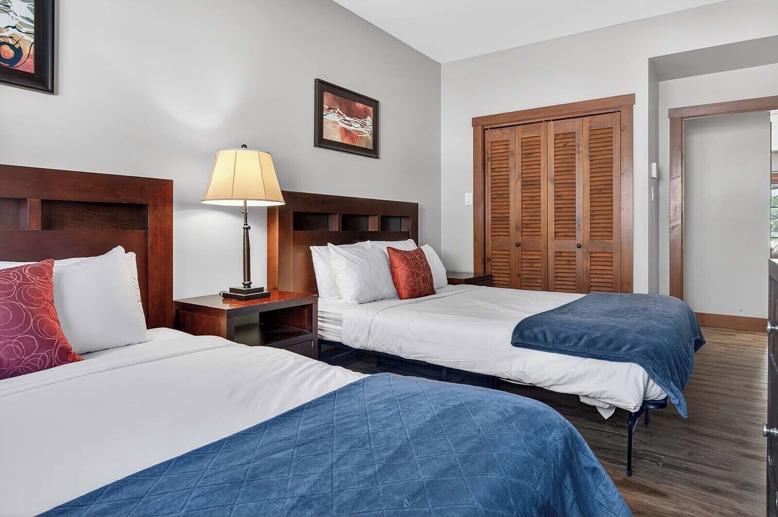 A hotel room with two beds, a nightstand with a lamp, a wooden wardrobe, and a framed picture on the wall.