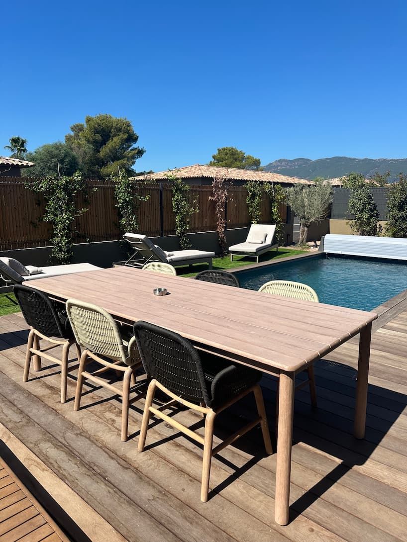 A wooden deck with a table and chairs, a swimming pool, and a fenced area with trees and a building in the background.