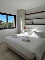 Una cama bien hecha con sábanas y toallas blancas, un ventanal con vistas a zonas verdes y un estante en la pared con un marco de fotos.