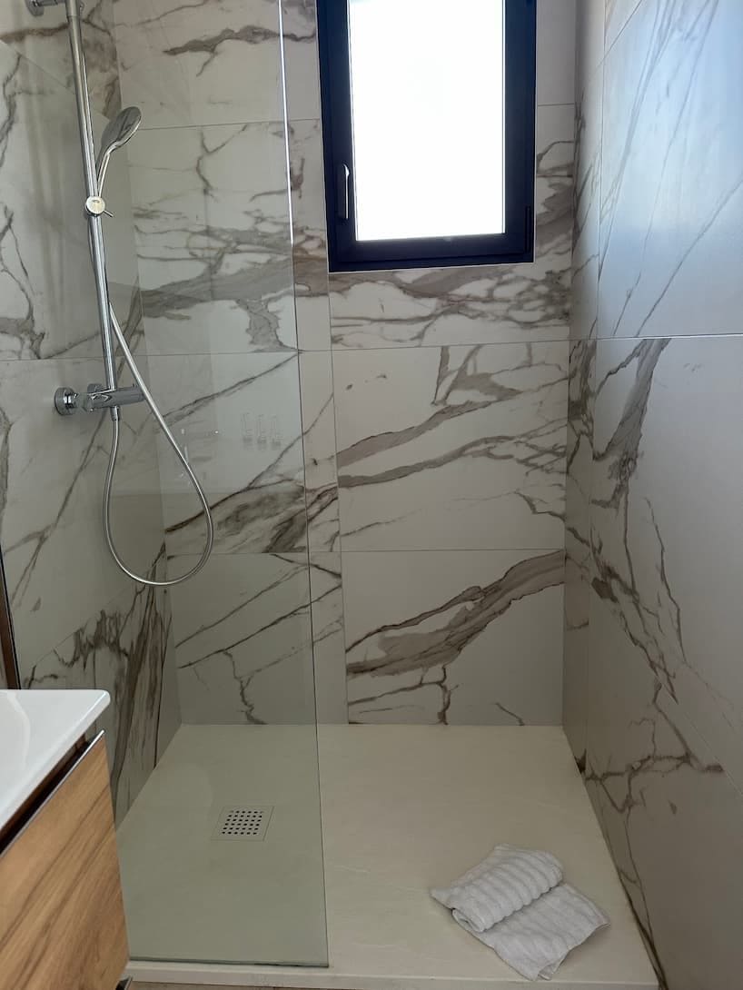 A bathroom with a glass shower enclosure, marble walls, a small window, and a towel on the floor.