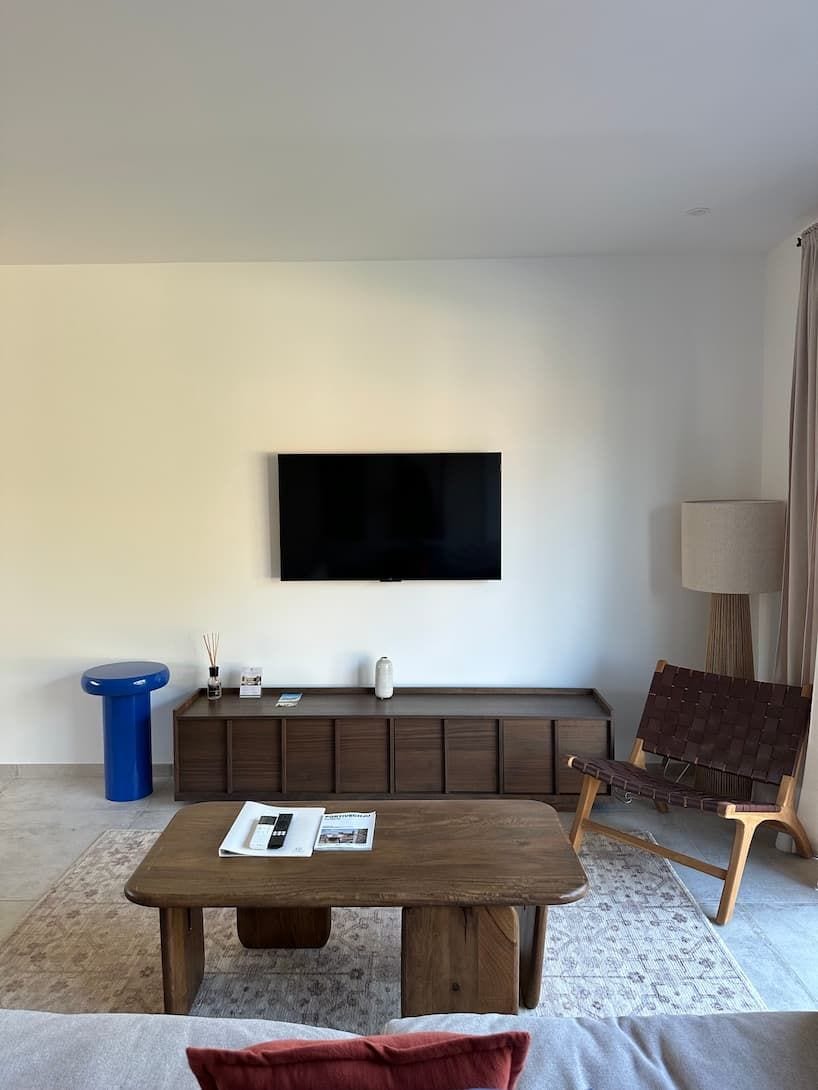 A modern living room with a flat-screen TV mounted on the wall, a wooden coffee table, a blue side table, a brown sofa, and a wicker chair.