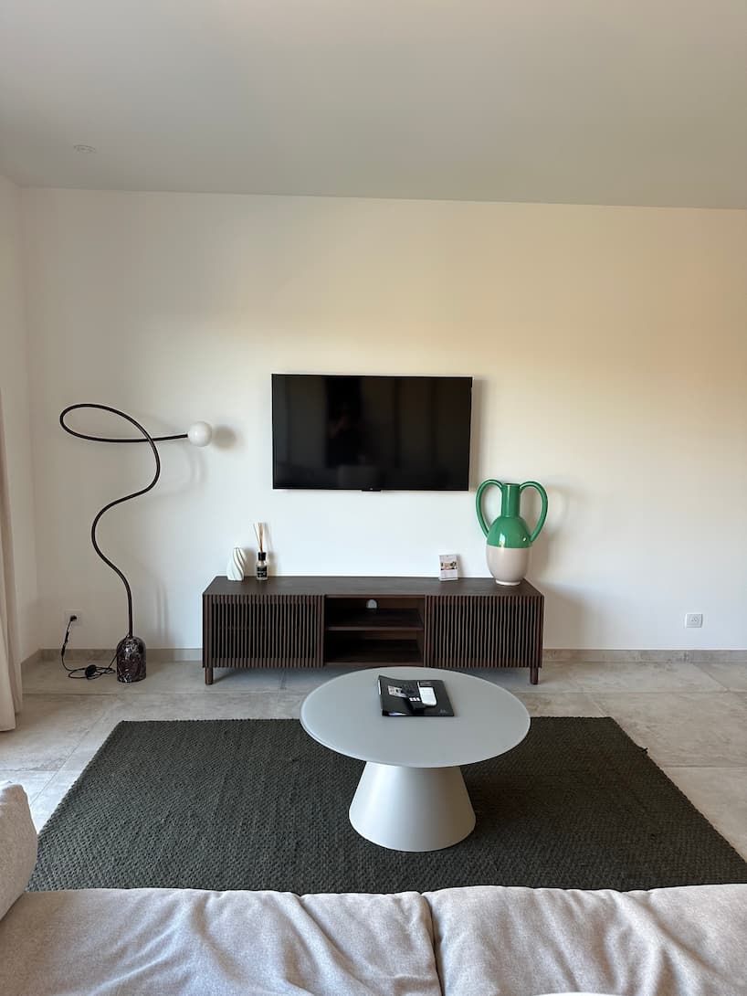 A modern living room with a flat-screen TV mounted on the wall, a dark wooden entertainment unit below, a round coffee table in the center, and a dark rug on the floor.