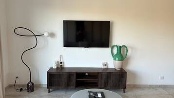 A modern living room with a flat-screen TV mounted on the wall, a dark wooden entertainment unit below, a round coffee table in the center, and a dark rug on the floor.