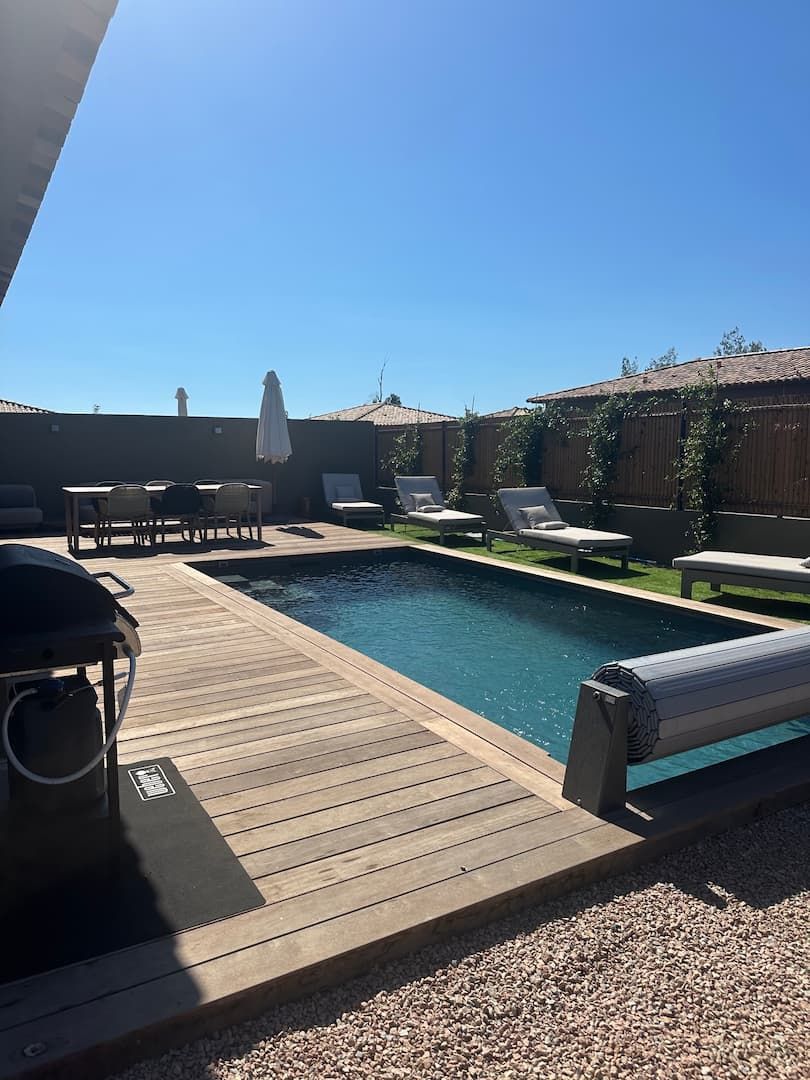A backyard with a swimming pool, wooden deck, lounge chairs, and a barbecue grill.
