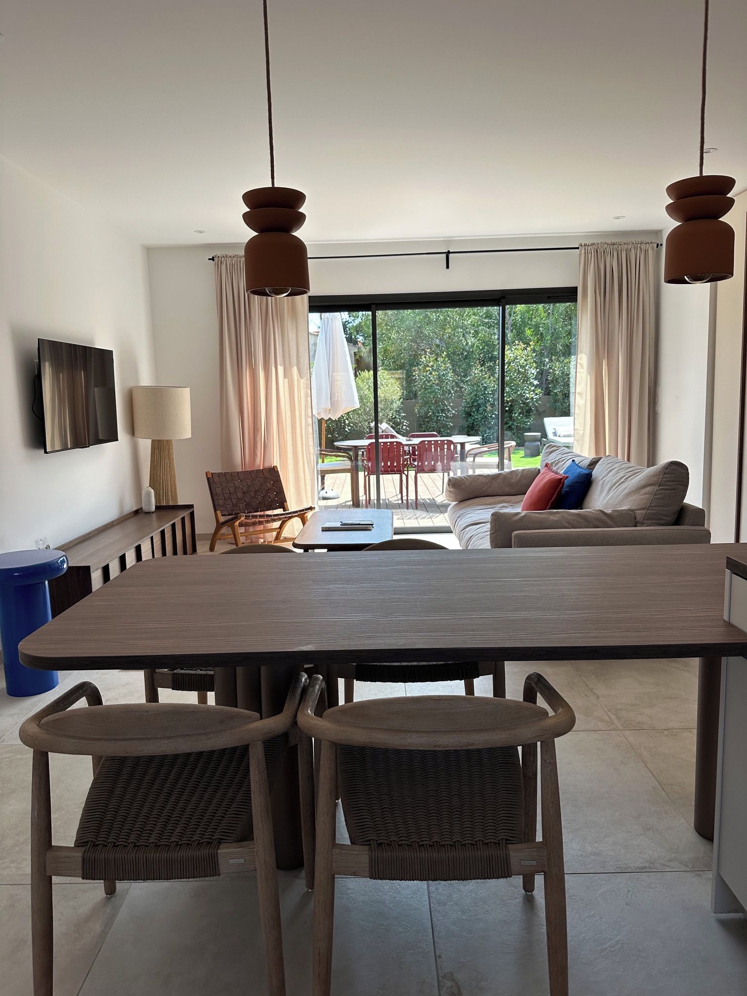 A modern living room with a large dining table, two wicker chairs, a sofa with cushions, a TV, and a window with curtains.