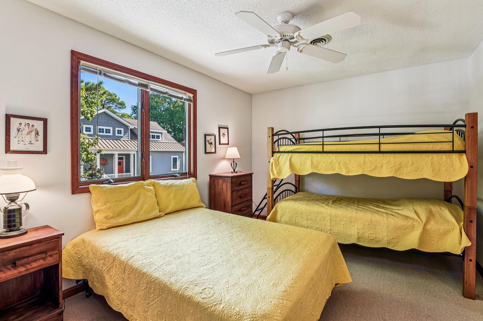 A room with a bunk bed, a desk, a lamp, a window with a view of houses, and a ceiling fan.