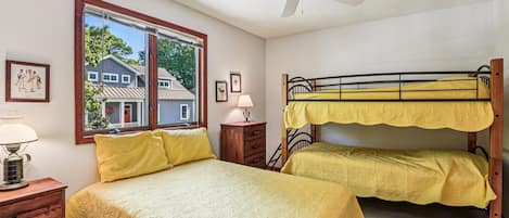 A room with a bunk bed, a desk, a lamp, a window with a view of houses, and a ceiling fan.