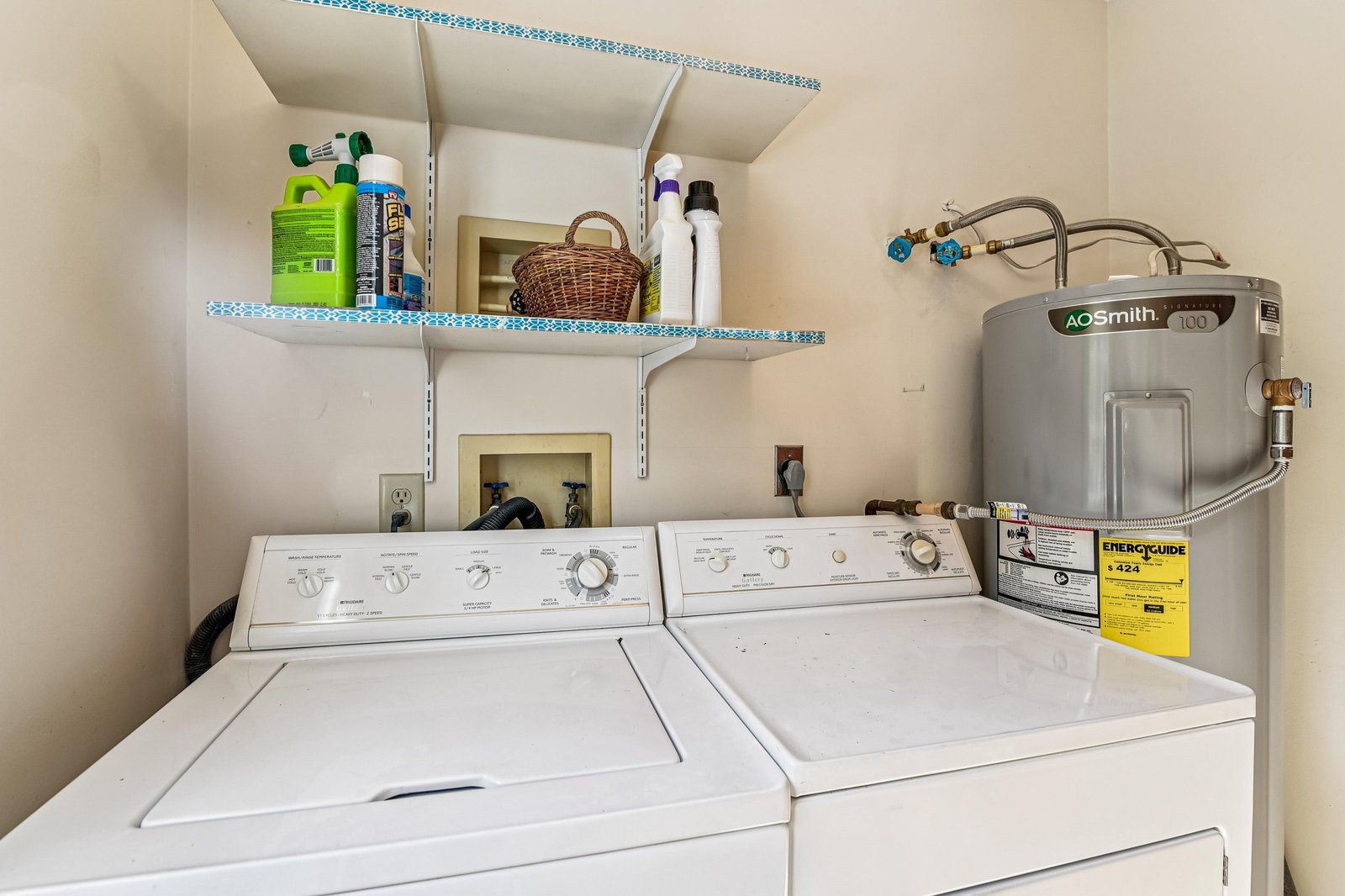 A laundry room with two white front-loading washers, a gray water heater, and shelves with cleaning supplies.