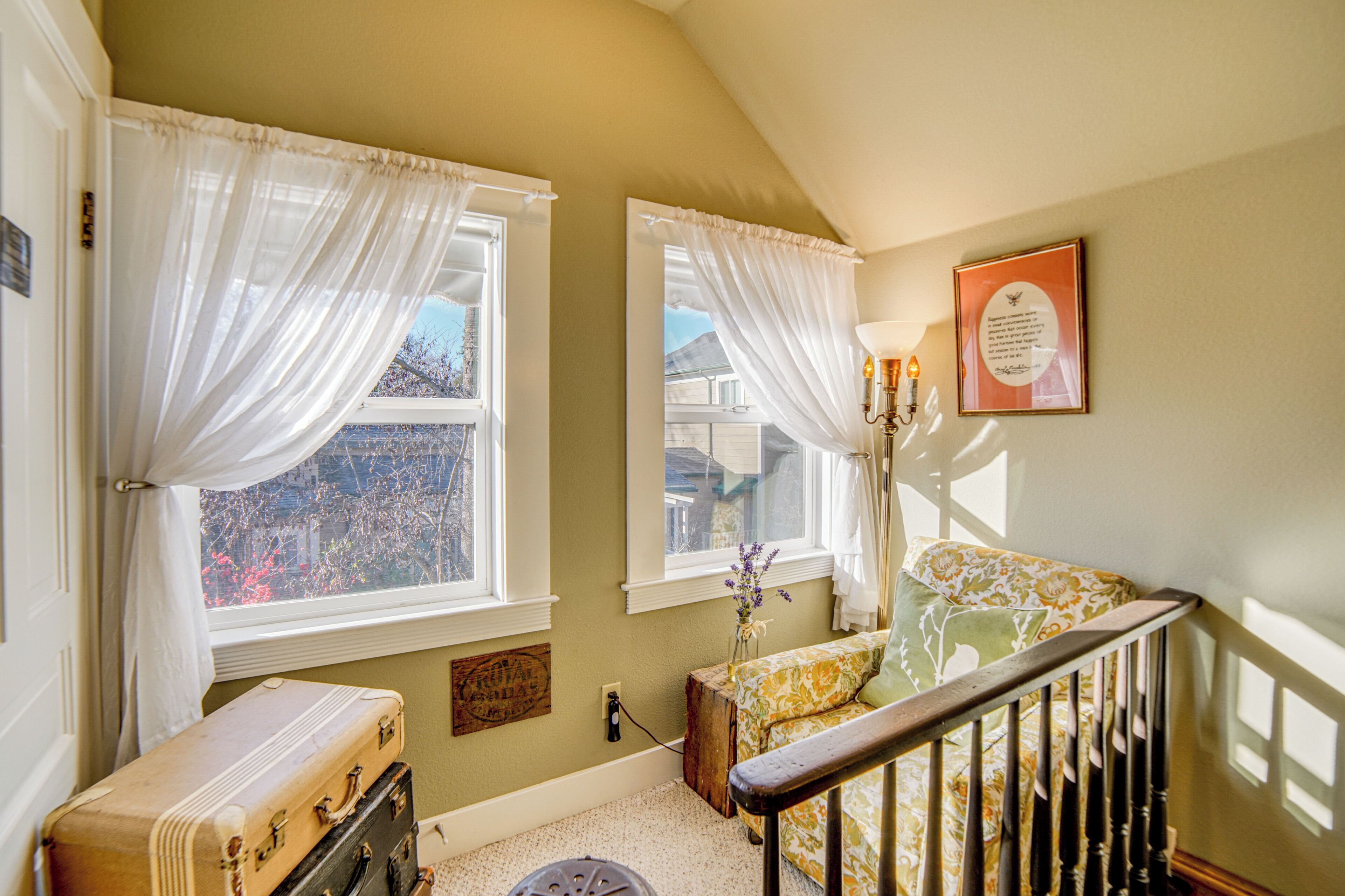 A room with two windows, a staircase, a vintage suitcase, and a framed quote on the wall.