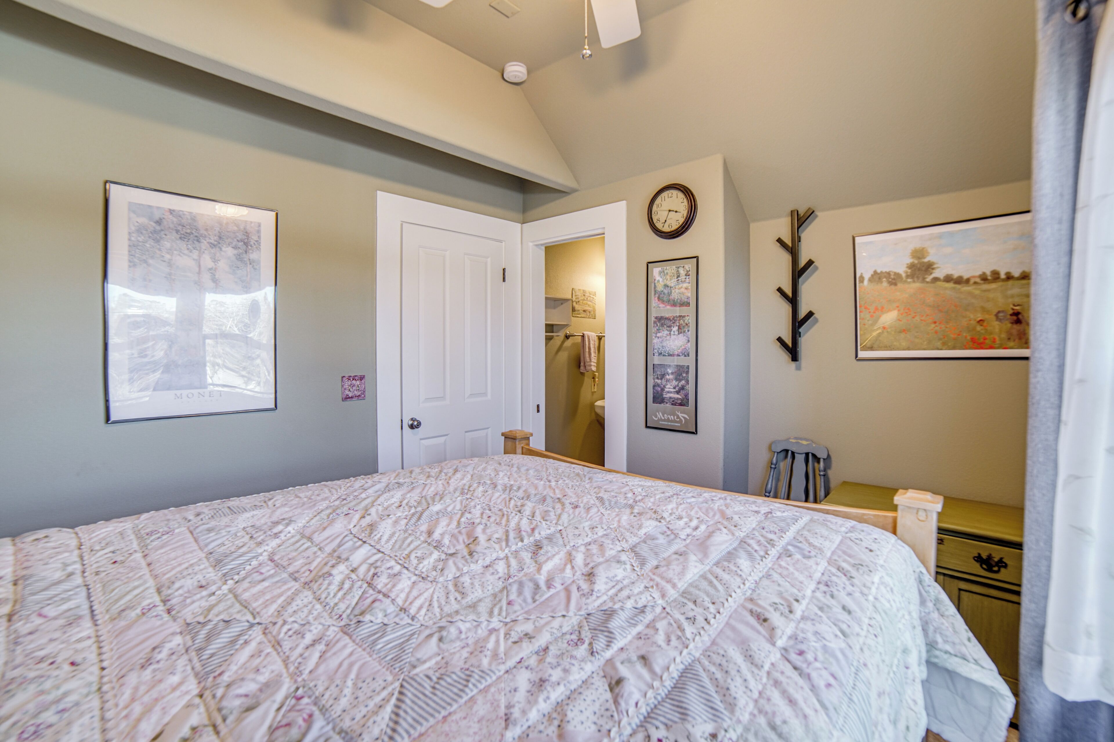 Un dormitorio con una cama grande, una puerta que da a un baño, un reloj en la pared y una pintura de paisaje.