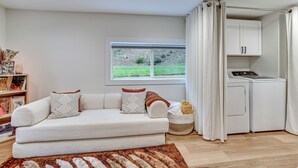 A modern living room with a sofa, a window with a view of greenery, a washing machine, and a bookshelf. - Walk to The Oyster Bar: Chuckanut Dr Retreat! (Bow)