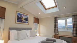 Une chambre avec un puits de lumière, un lit avec des oreillers, une table de chevet avec une lampe, un bureau et une fenêtre avec des rideaux.